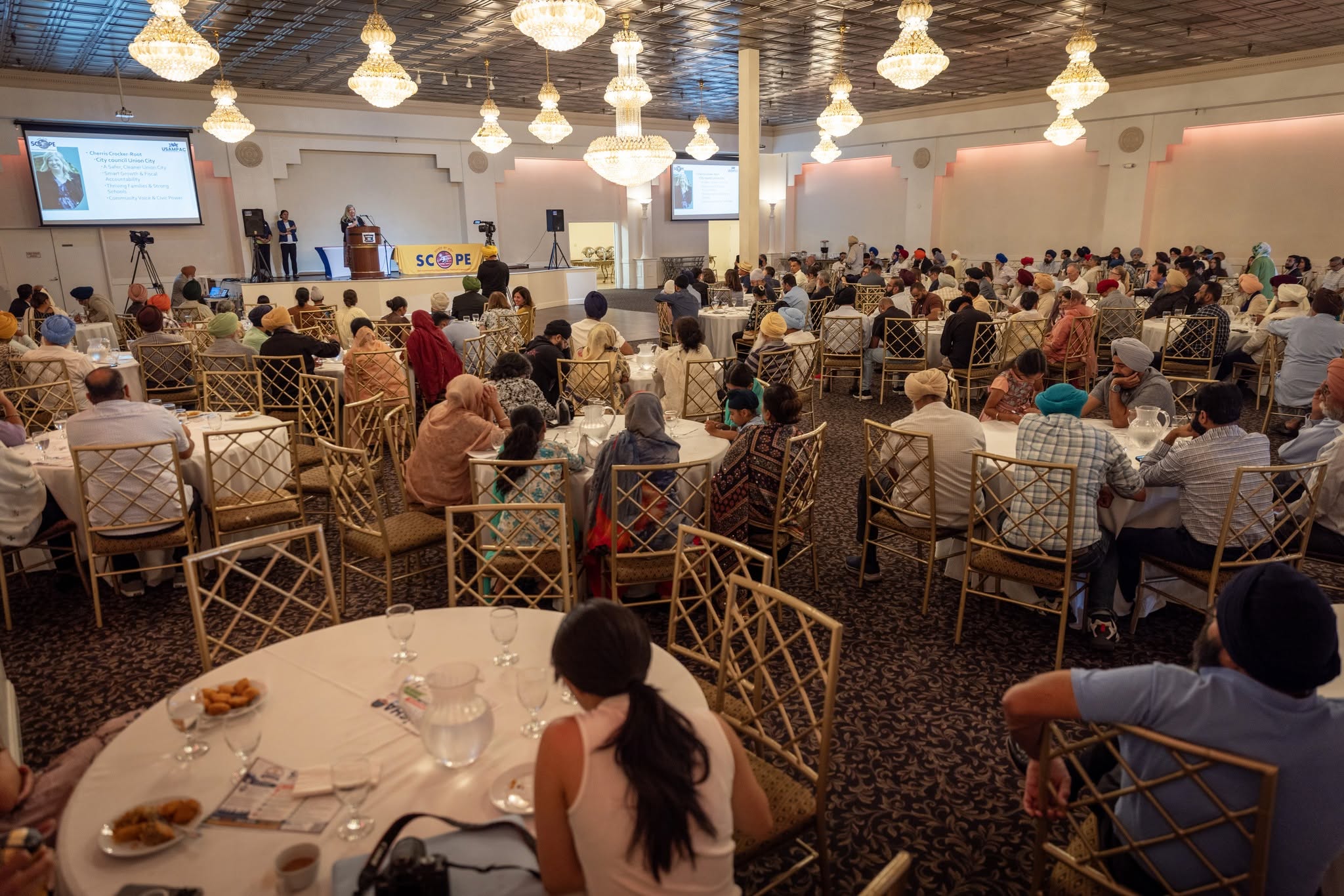 Guests seated at round tables in a chandelier-lit banquet hall watch a speaker at a podium beside a SCOPE banner and projection screens
